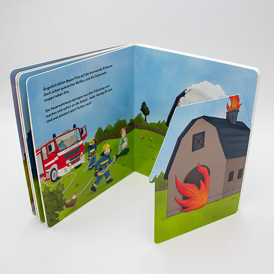 Das Buchcover zeigt einen brennenden Stall mit roten Flammen, ein rotes Löschfahrzeug und Feuerwehrleute in blauen Uniformen auf grüner Wiese.