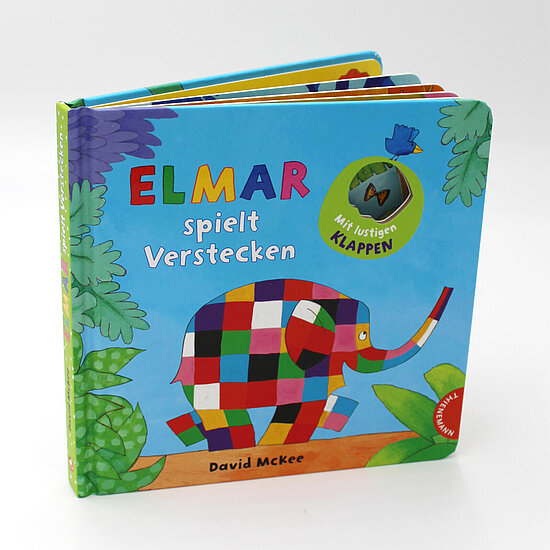 Das Buchcover zeigt Elmar, den bunt karierten Elefanten, vor hellblauem Hintergrund mit grünen Blättern, kleinem blauen Vogel und orangem Boden.