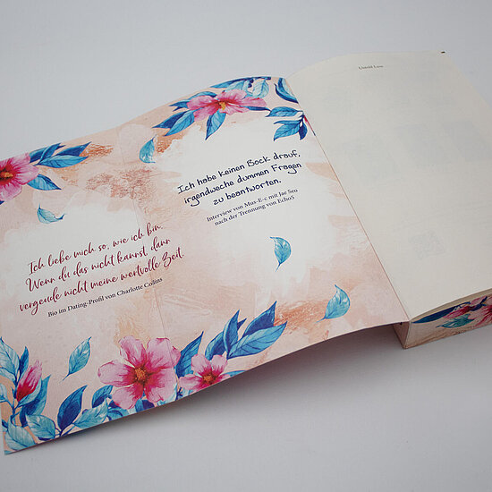Das Buchcover zeigt ein offenes Buch mit aquarellierten rosa Blüten und blauen Blättern auf hellem pfirsichfarbenem Hintergrund.