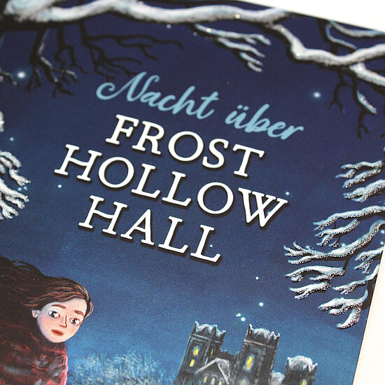 Das Buchcover zeigt einen dunkelblauen Nachthimmel mit schneebesetzten Ästen, links ein Mädchen im roten Mantel und unten ein beleuchtetes Herrenhaus.