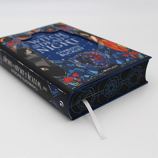 Das Buchcover zeigt … eine blaurote glasmalereiartige Illustration mit einer Frau im schwarzen Umhang, roten Rosen und einem magischen Kreis.