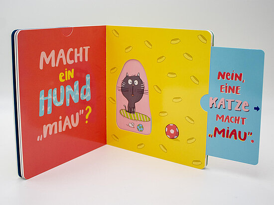 Das Buchcover zeigt … ein aufgeschlagenes Buch in Rot Gelb und Blau mit einem schwarzen Kater in einer rosa Aussparung sowie Spielball und Muster.