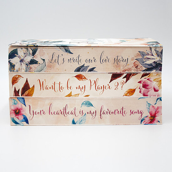 Das Buchcover zeigt drei gestapelte Buchrücken mit floralen Aquarellblüten in Pastellrosa, Pastellblau und Beige sowie eleganter Handschrift.