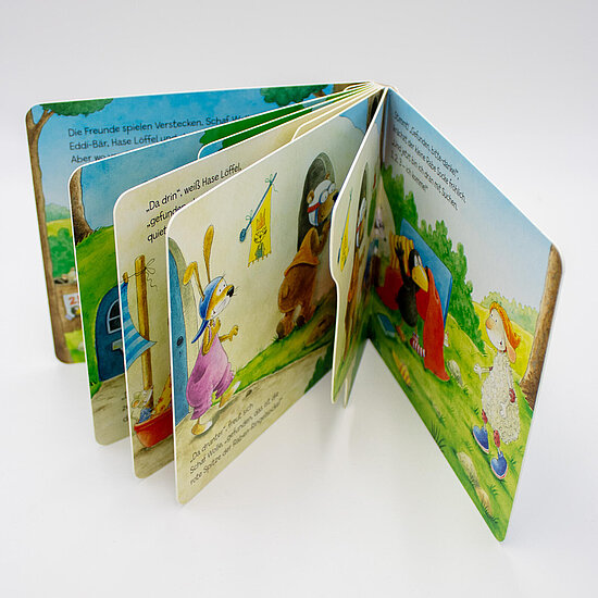 Das Buchcover zeigt ein aufgeklapptes Pappbilderbuch mit bunten Tierfiguren, Hase und Schafe, vor grünem Wiesenhintergrund.