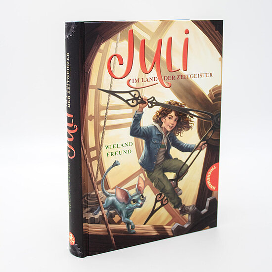 Das Buchcover zeigt … ein Mädchen mit lockigen Haaren und Jeansjacke an einem großen Uhrzeiger hängend, daneben eine blaue Katze vor goldenem Uhrwerk.