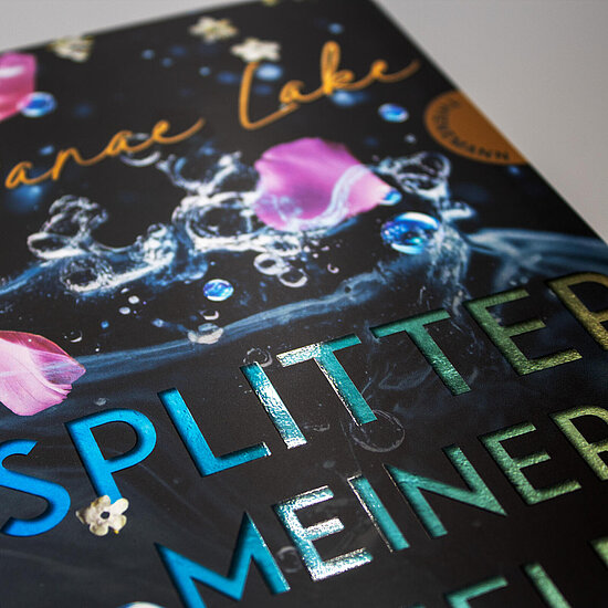 Das Buchcover zeigt vor schwarzem Grund eine Wasserfontäne mit Spritzern und blauen Luftblasen, rosa Blütenblättern und blaugrün schimmerndem Titel.