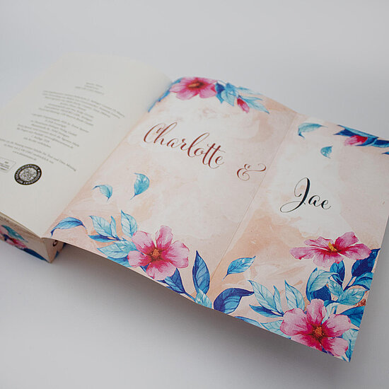 Das Buchcover zeigt einen offenen Einband mit pastellbeigem Hintergrund, kalligrafischen Namen und pinken Blumen sowie blauen Blättern.